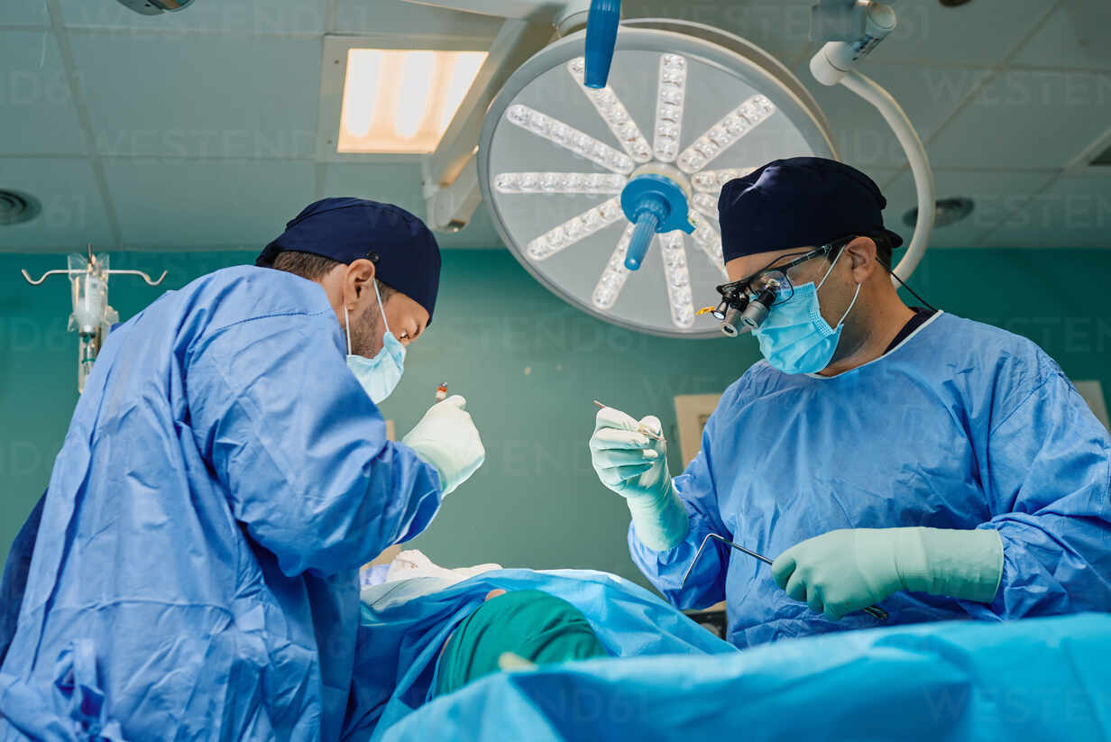 Unrecognizable patient lying on operating table during surgery performing by concentrated male doctors in medical gowns and masks